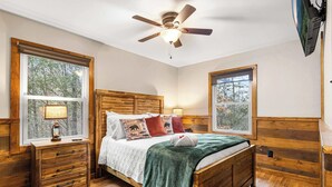Un dormitorio con cama de madera, dos mesitas de noche, ventilador de techo y ventanas amplias con vistas a árboles.