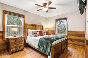 A bedroom with a wooden bed, two bedside tables, a ceiling fan, and large windows with views of trees.
