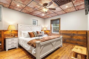 Un dormitorio con cabecera de madera, una cama con varias almohadas, mesitas de noche con lámparas, un ventilador de techo y una ventana con vistas a árboles.