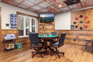A pool table with chairs, a dartboard, and a view of a forest through a window.