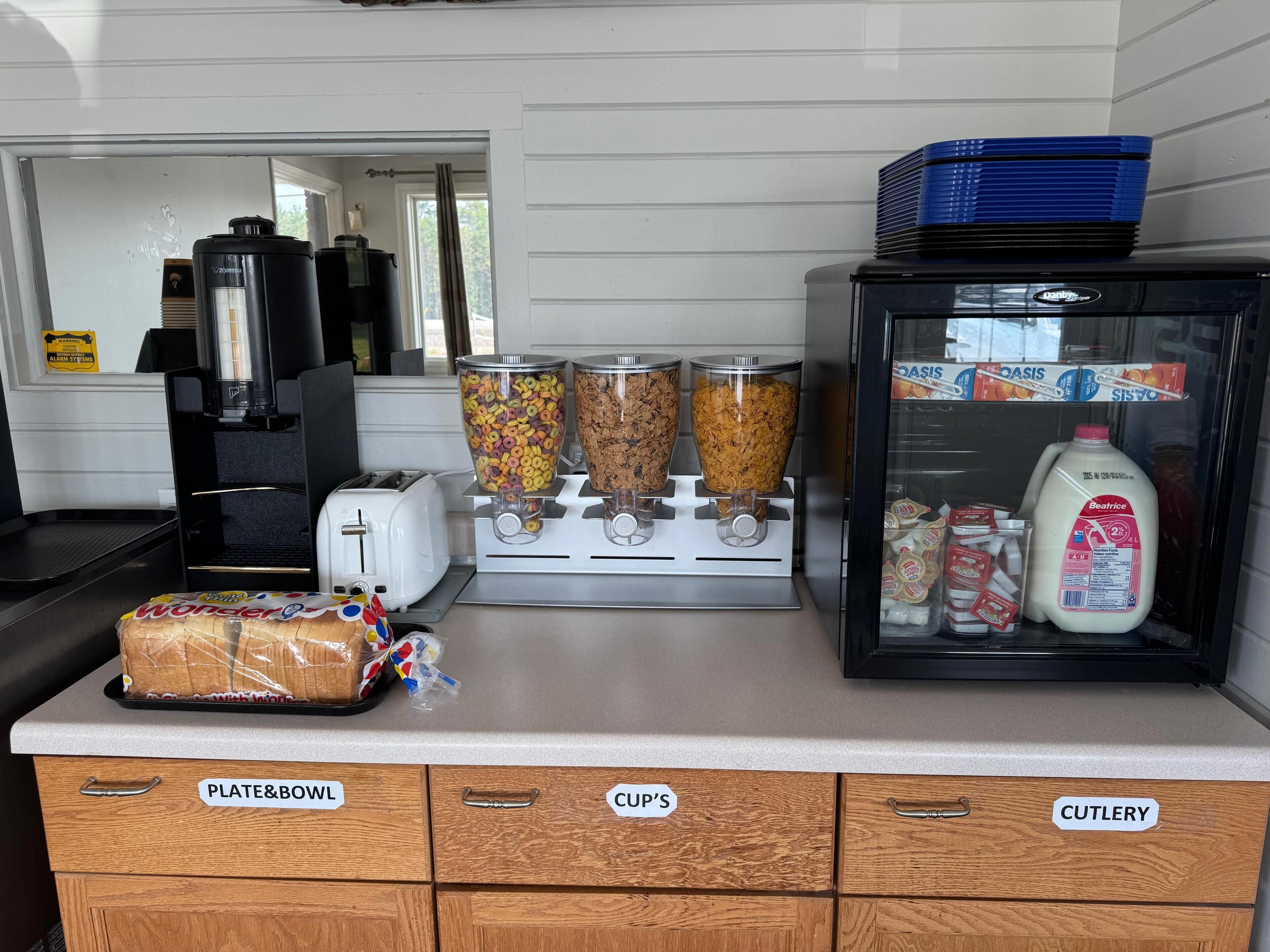 Un espace cuisine comprenant un présentoir à pain, des distributeurs de céréales, un réfrigérateur à lait et des tiroirs étiquetés pour les assiettes et les bols, les tasses et les couverts.