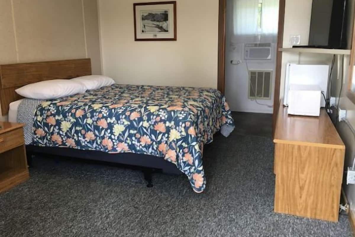 A hotel room with a bed, a wooden nightstand, a desk with a television, and a door leading to a bathroom.
