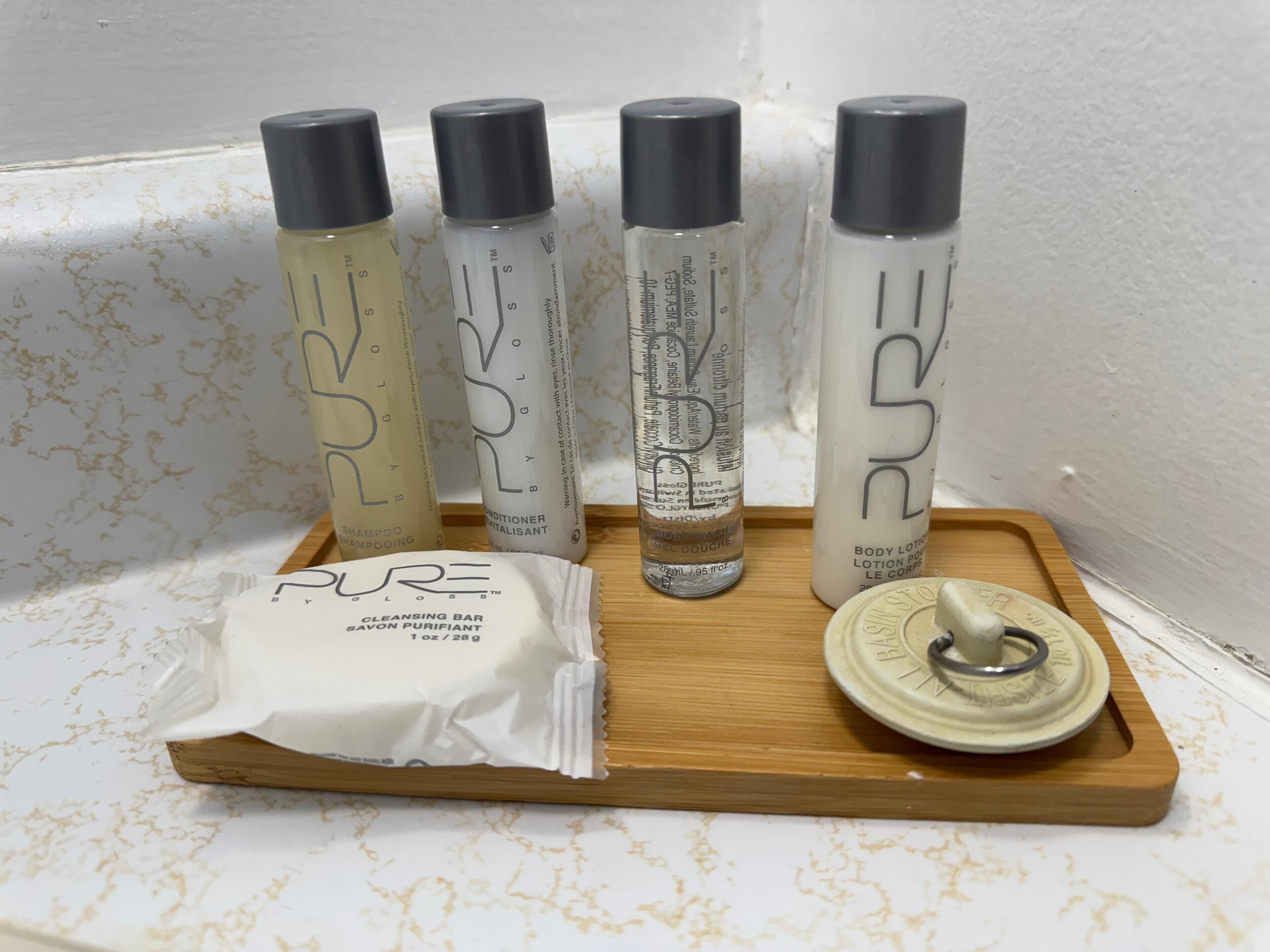 A wooden tray with four bottles of PURE body care products and a sachet of PURE cleansing bar on a white textured surface.