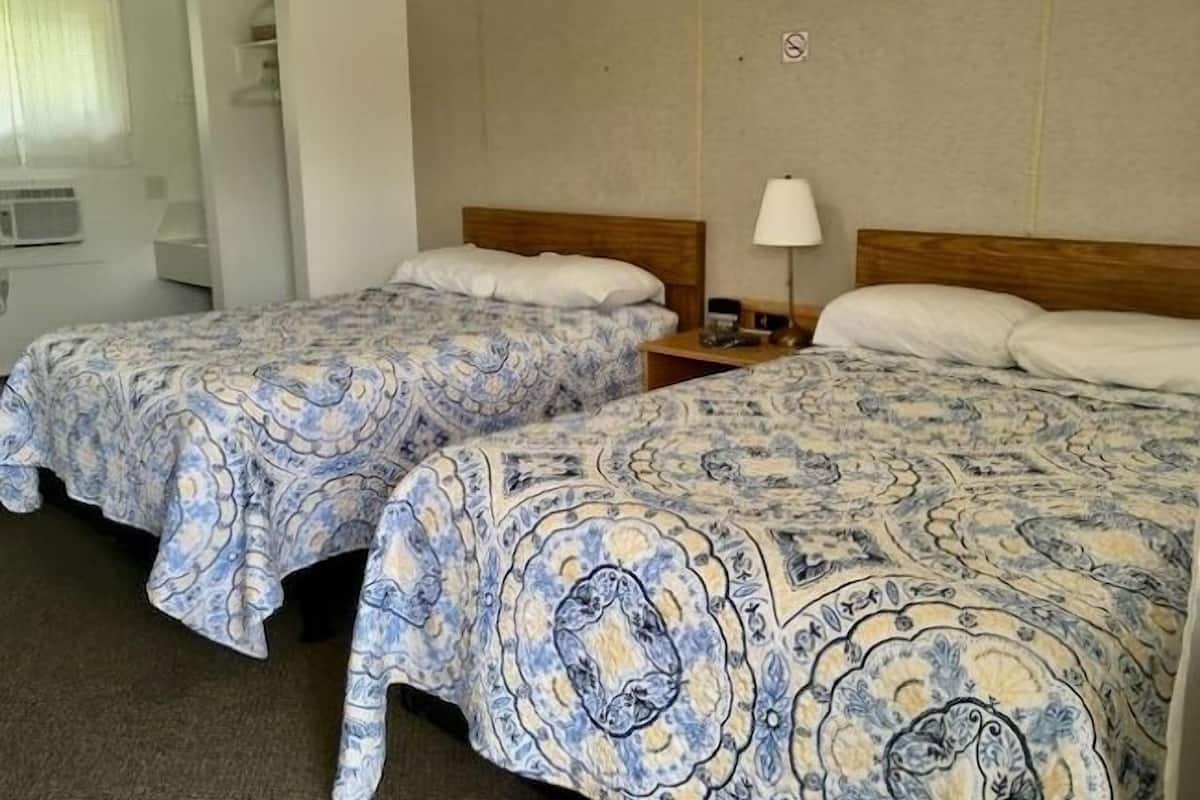 Two single beds with patterned bedding, a bedside table with a lamp, and a wall-mounted shelf.