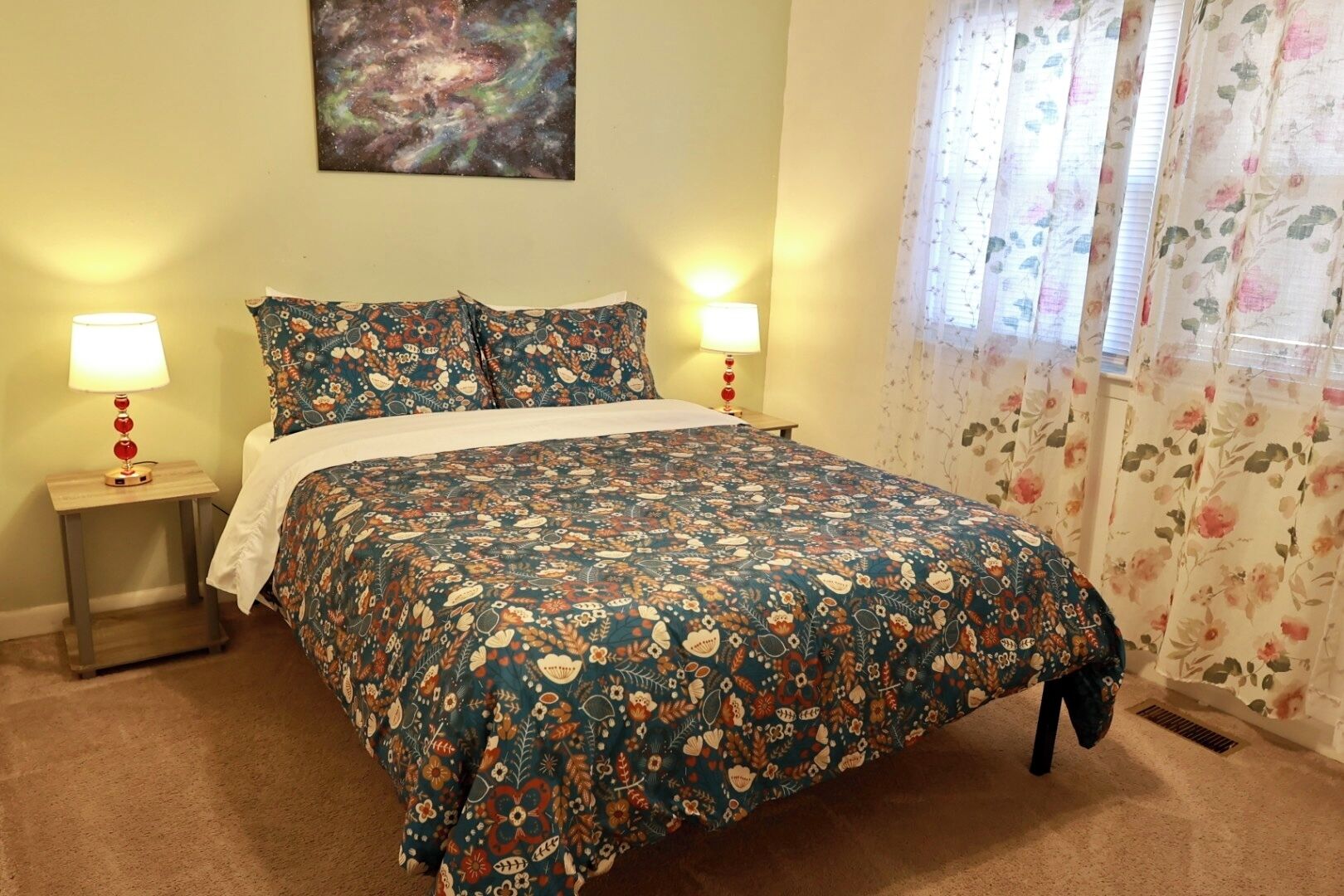 A bedroom with a floral-patterned bedspread, two bedside lamps, a galaxy-themed wall art, and floral curtains.