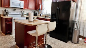 A kitchen with wooden cabinets, a black refrigerator, a microwave, a coffee maker, and a bar stool. - Cozy, gallery-inspired stay, 5min to Fort Liberty (Fayetteville)