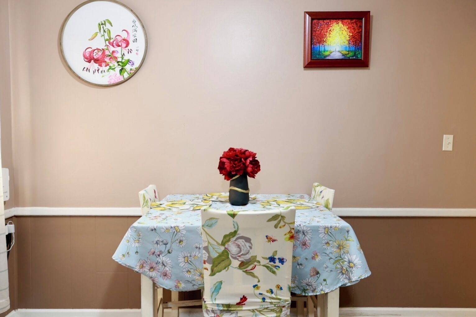 A dining area with a table covered in floral tablecloths, a vase of red flowers, and two framed artworks on the wall.