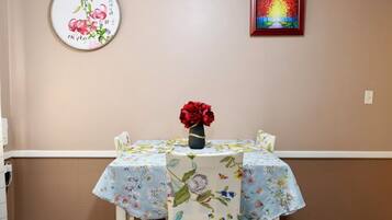A dining area with a table covered in floral tablecloths, a vase of red flowers, and two framed artworks on the wall.