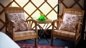 Zwei Korbstühle mit floralen Kissen, ein kleiner runder Tisch mit einem grünen Krug und Gläsern, und Blick durch ein Gitterfenster.