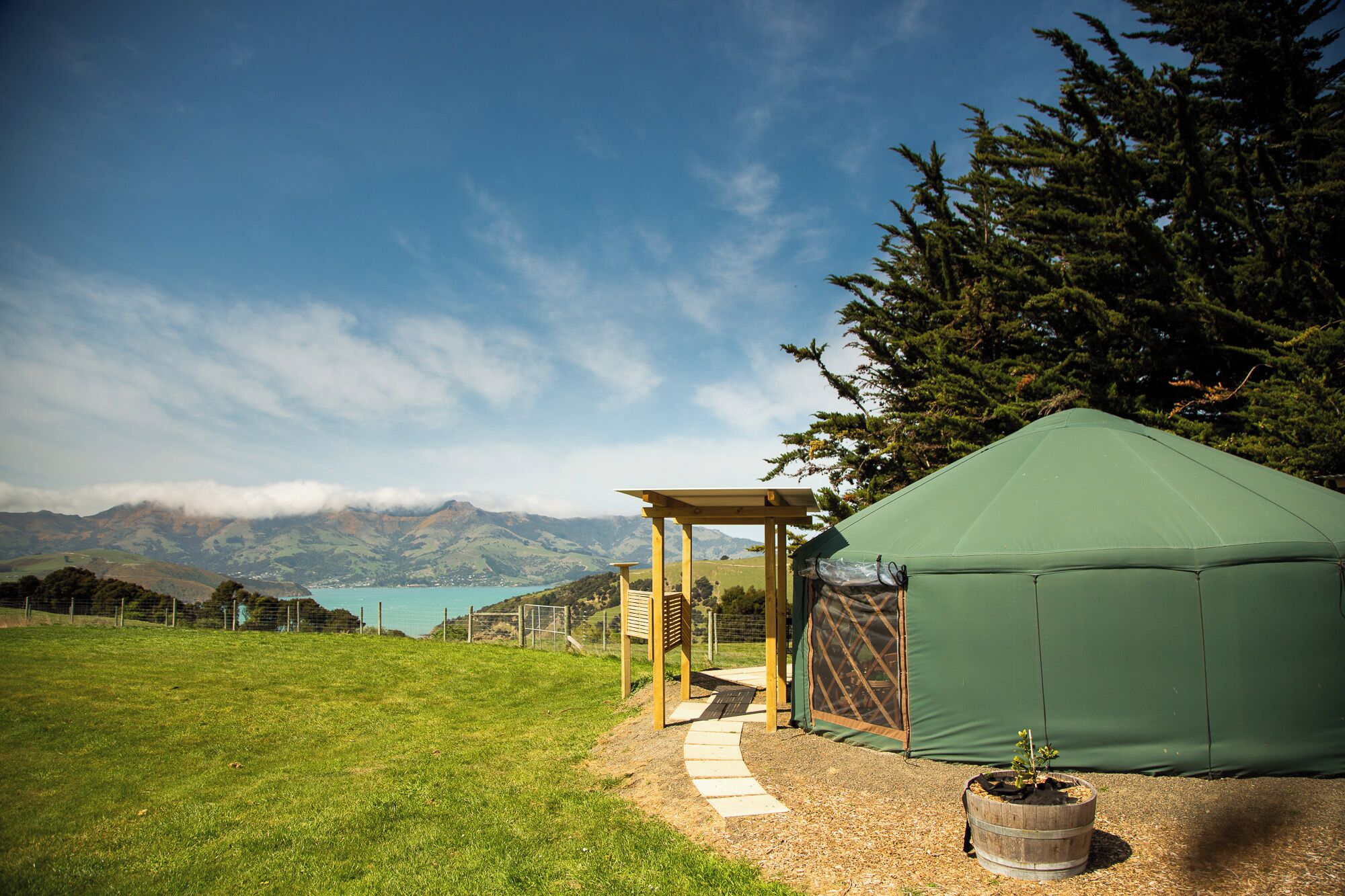 A green tent-like structure with a wooden pergola entrance, a small plant in a barrel, and a clear view of distant mountains and a lake.