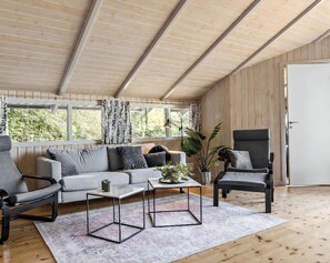 A modern living room with a wooden ceiling, a sofa, two armchairs, a coffee table, and a view of greenery through large windows.
