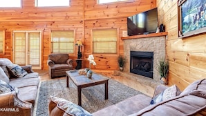 A cozy log cabin interior with a fireplace, leather sofas, and a flat-screen TV mounted above it.