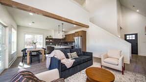 A spacious living area with a dark grey sofa, a white armchair, and a wooden coffee table. A dining table with chairs is positioned near a large window.