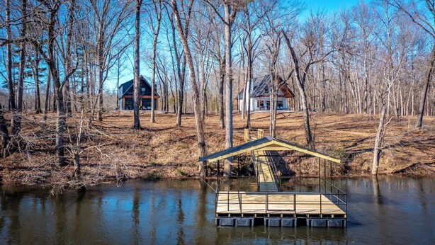 A wooden dock extending into a calm body of water, surrounded by leafless trees and two houses in the background.