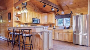 A wooden cabin kitchen with a central island, bar stools, a stainless steel refrigerator, and a window with a view of the outdoors.