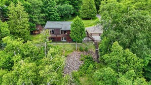A cabin surrounded by dense forest, with a wooden deck and a staircase leading to a porch.