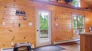 A wooden cabin interior with a door leading outside, a small shelf with decorative items, and a pet feeding station.
