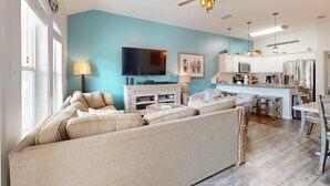 Ein Wohnzimmer mit beigefarbenem Sofa, Fernseher und Küchenzeile mit weißen Schränken und Holzboden.