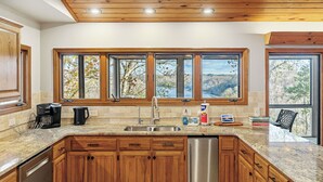 A kitchen with wooden cabinets, a stainless steel dishwasher, a coffee maker, and a window with a view of trees and mountains.