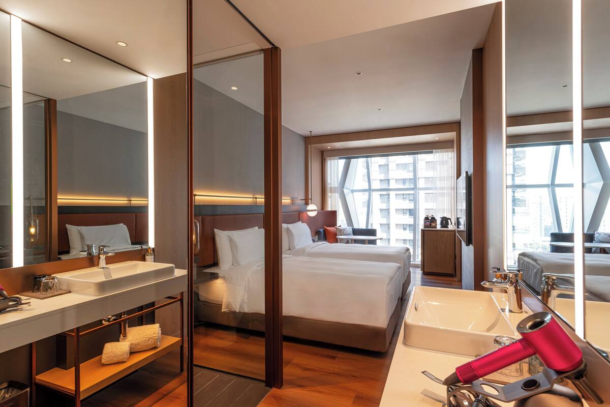 A modern hotel room with a large bed, a separate bathroom with a sink and mirror, and a view of the city through large windows.