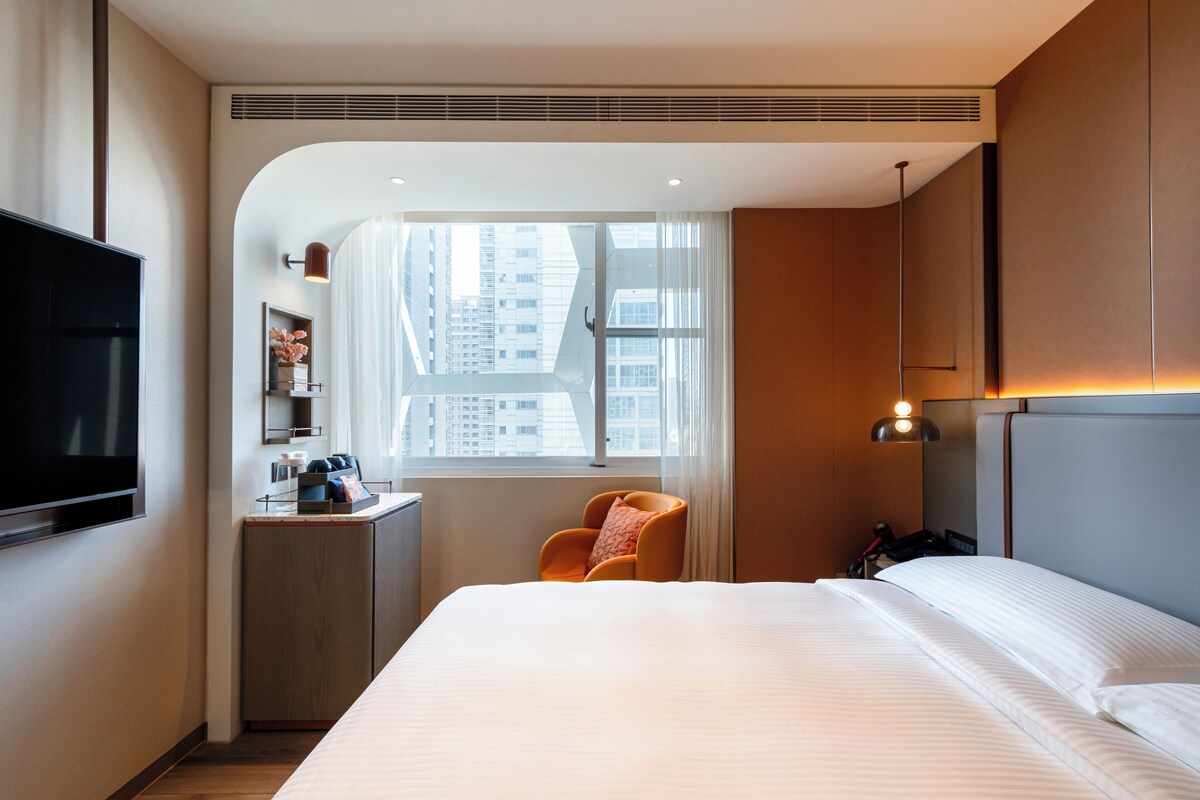 A hotel room with a large bed, a TV, a desk with a chair, a window with a view of buildings, and a lamp.