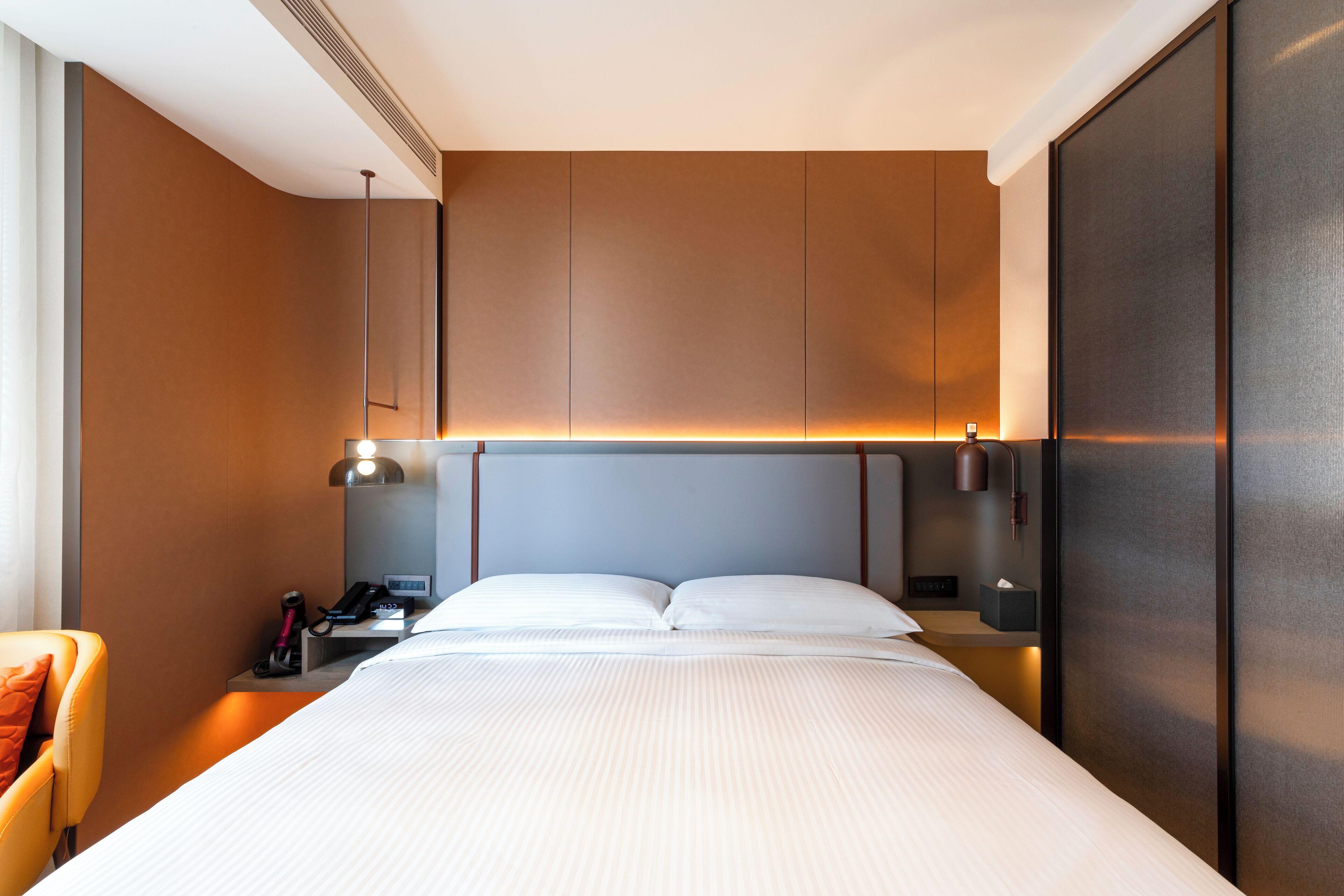 A modern hotel room with a large bed, a nightstand with a lamp, a chair, and a wardrobe.