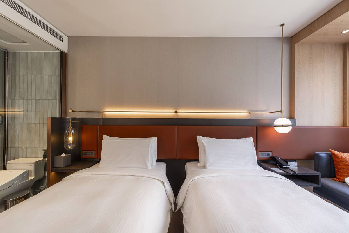 A hotel room with two beds, a bedside table with a lamp, a small table with a phone, and a window with blinds.