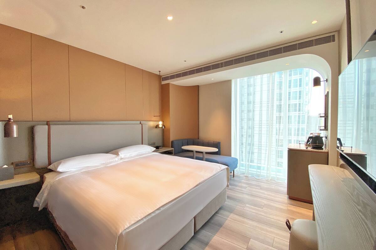 A hotel room with a large bed, a seating area, and a view of the city through large windows.