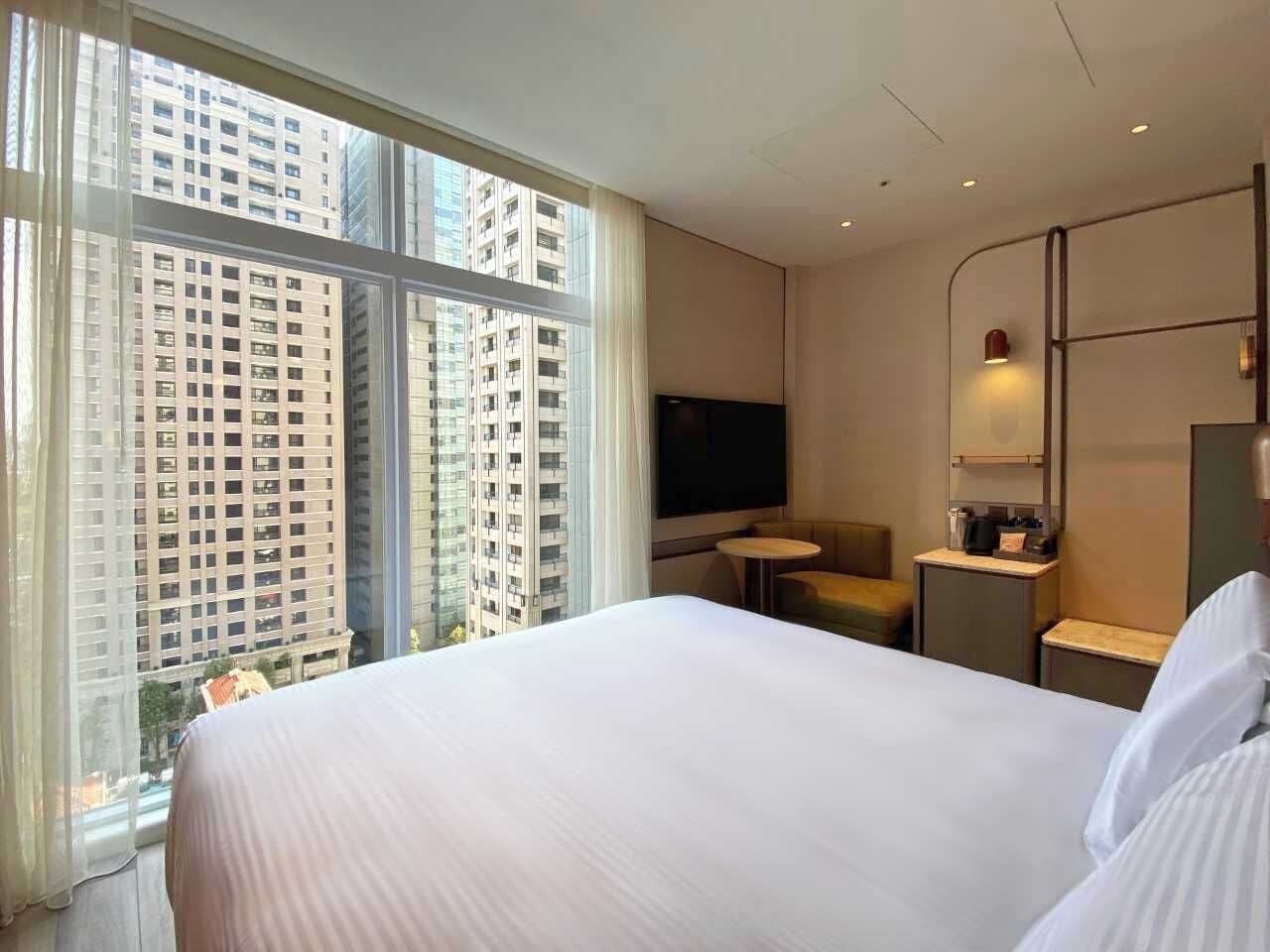 A hotel room with a large bed, a TV, a small table, a chair, and a view of tall buildings.