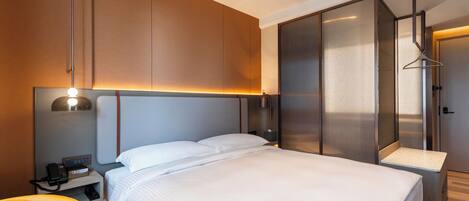 A modern hotel room with a large bed, a bedside table with a lamp, a chair, and a wardrobe.