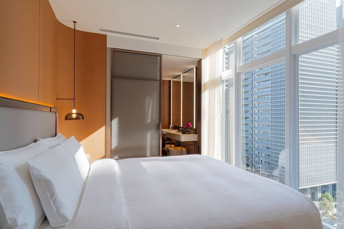 A modern hotel room with a large bed, a desk, and a view of a cityscape through large windows.