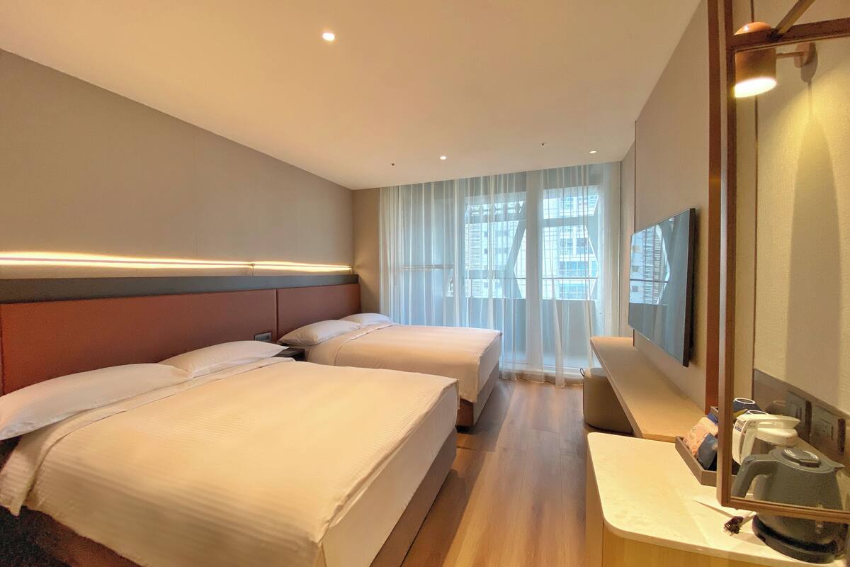 A hotel room with two beds, a large mirror, and a view of the city through sheer curtains.