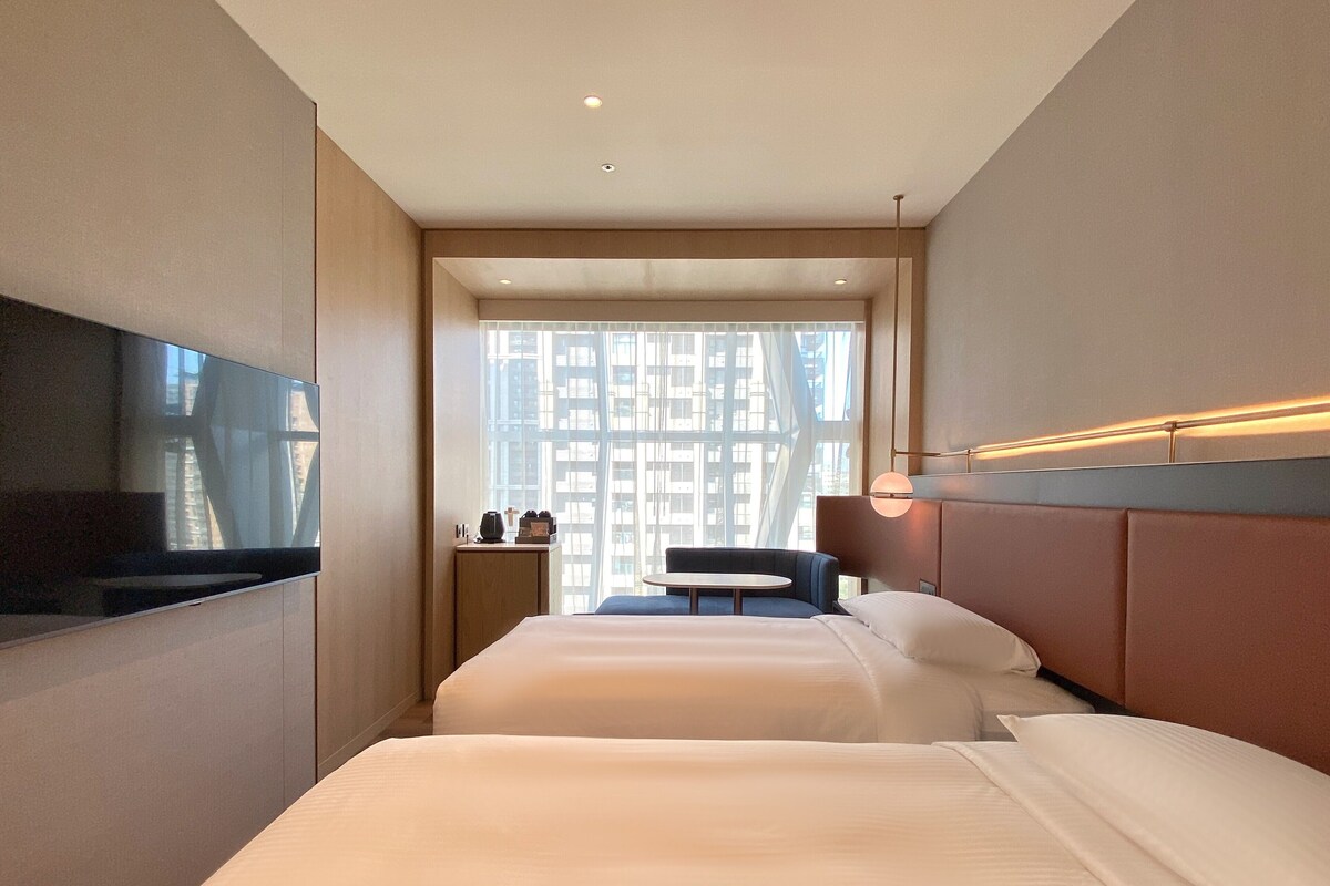 A modern hotel room with a large bed, a flat-screen TV, a small table, and a view of a cityscape through large windows.