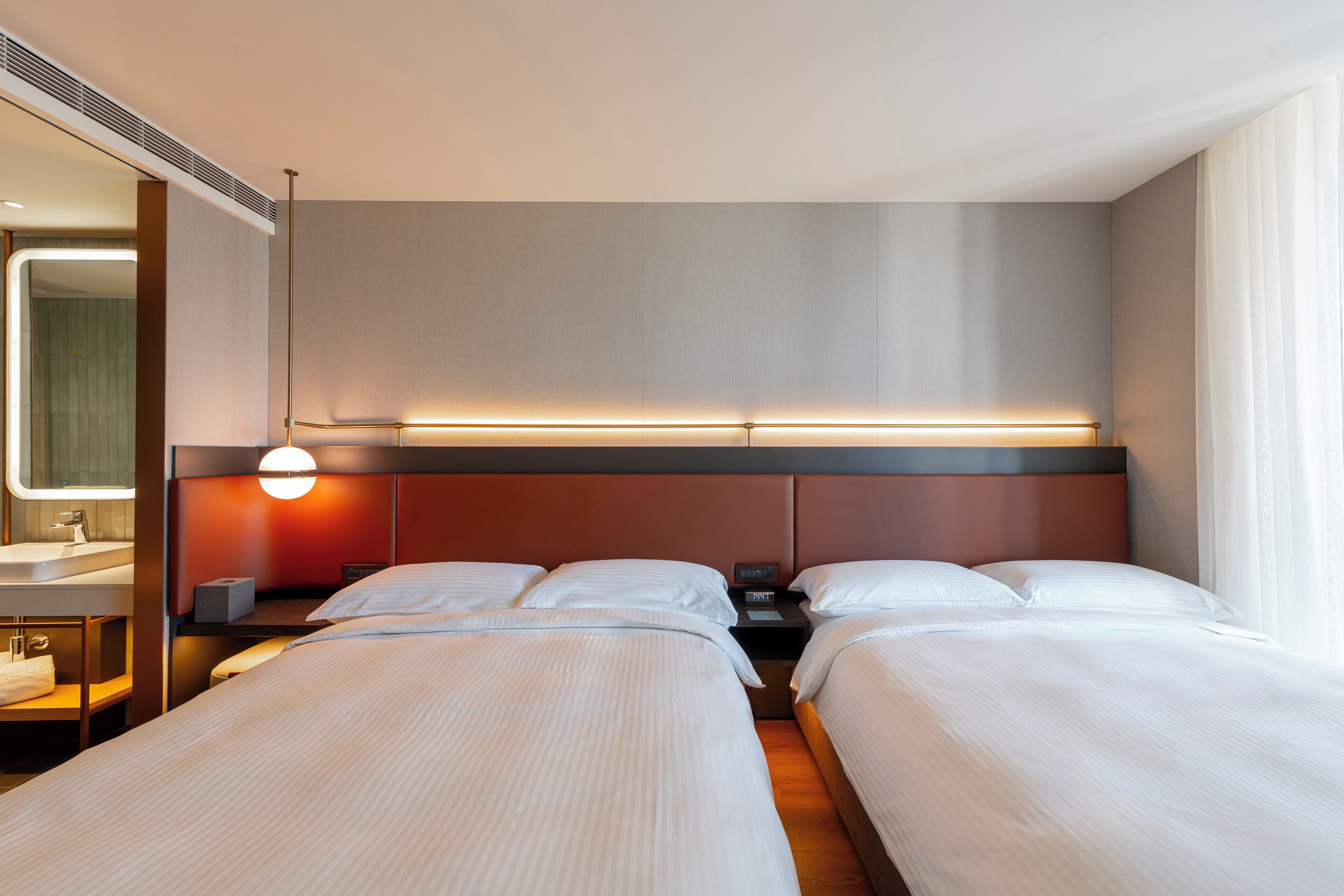 Hotelkamer met twee bedden, een nachtkastje met lamp, een spiegel en een badkamer met wastafel en handdoekrek.