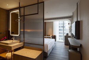 Habitación de hotel moderna con una cama grande, una zona de estar, un baño con tocador y un ventanal con vistas a la ciudad.