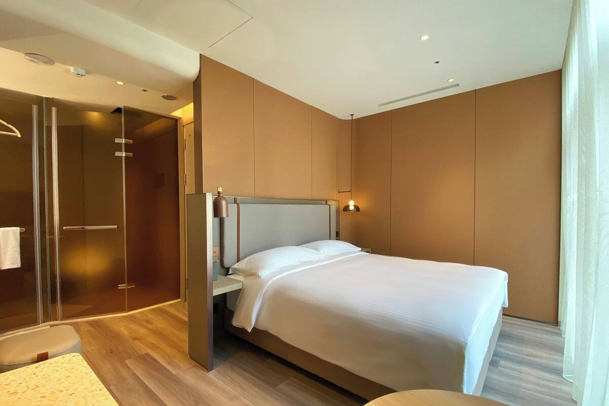 A modern hotel room with a bed, a glass shower enclosure, and a wooden floor.