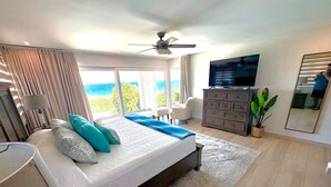 Une chambre avec un grand lit, un ventilateur de plafond, une télévision, une commode et une vue sur l’océan.