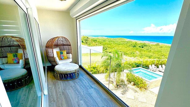 Un balcon agrémenté de fauteuils en osier et d’un tambour, donnant sur une piscine et une végétation luxuriante.
