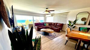 Un salon spacieux avec un grand canapé, une table basse en bois et une table à manger. Il y a un ventilateur de plafond et une vue sur l’océan par de grandes fenêtres.