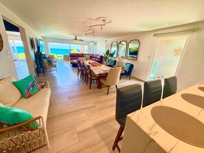Een ruime woonkamer met een grote eethoek, een bank met groene kussens en uitzicht op de oceaan door grote ramen.