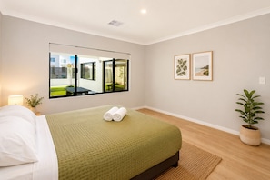 A hotel room with a bed, a window with a view of a building, two framed pictures on the wall, and a potted plant. - 5 O Clock Somewhere (West Busselton)