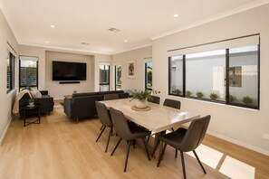 A modern living room with a sofa set, a dining table with chairs, a flat-screen TV mounted on the wall, and large windows with a view of the outside. - 5 O Clock Somewhere (West Busselton)