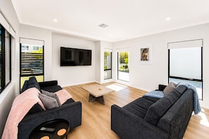A modern living room with a flat-screen TV, two sofas, a coffee table, and large windows with blinds. - 5 O Clock Somewhere (West Busselton)
