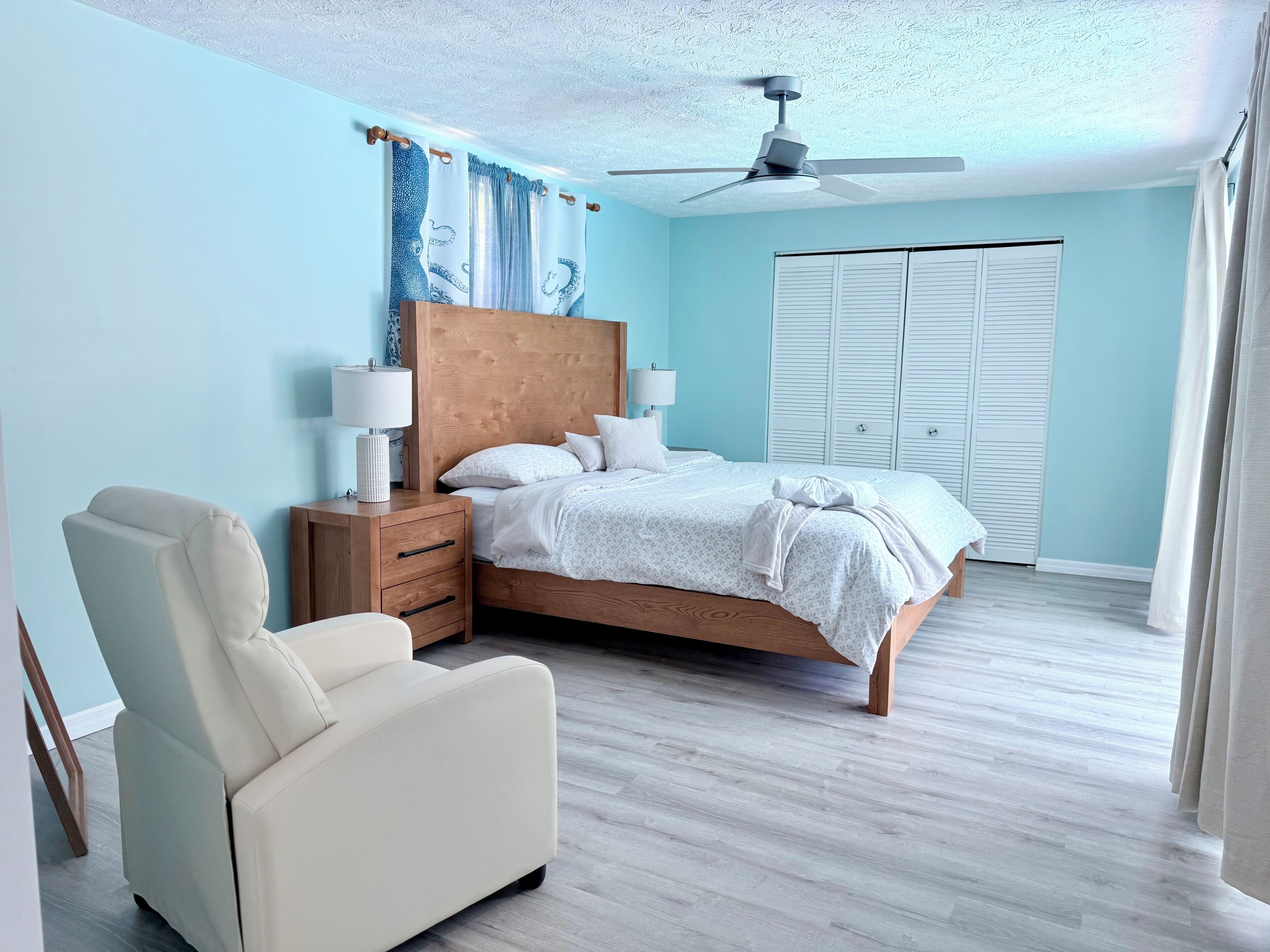 A bedroom with a bed, nightstands, a chair, a ceiling fan, and a window with shutters.