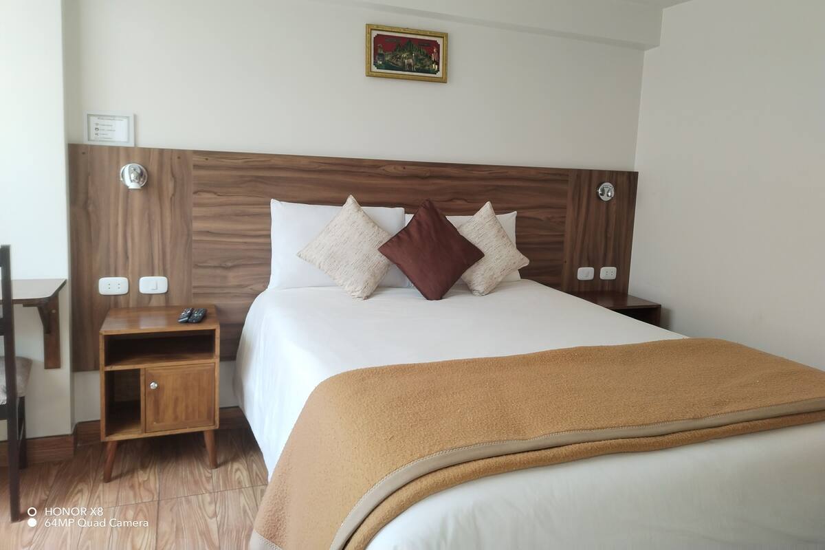 A hotel room with a wooden headboard, a bed with white linens, two pillows, a brown throw blanket, a nightstand with a phone, and a small table.