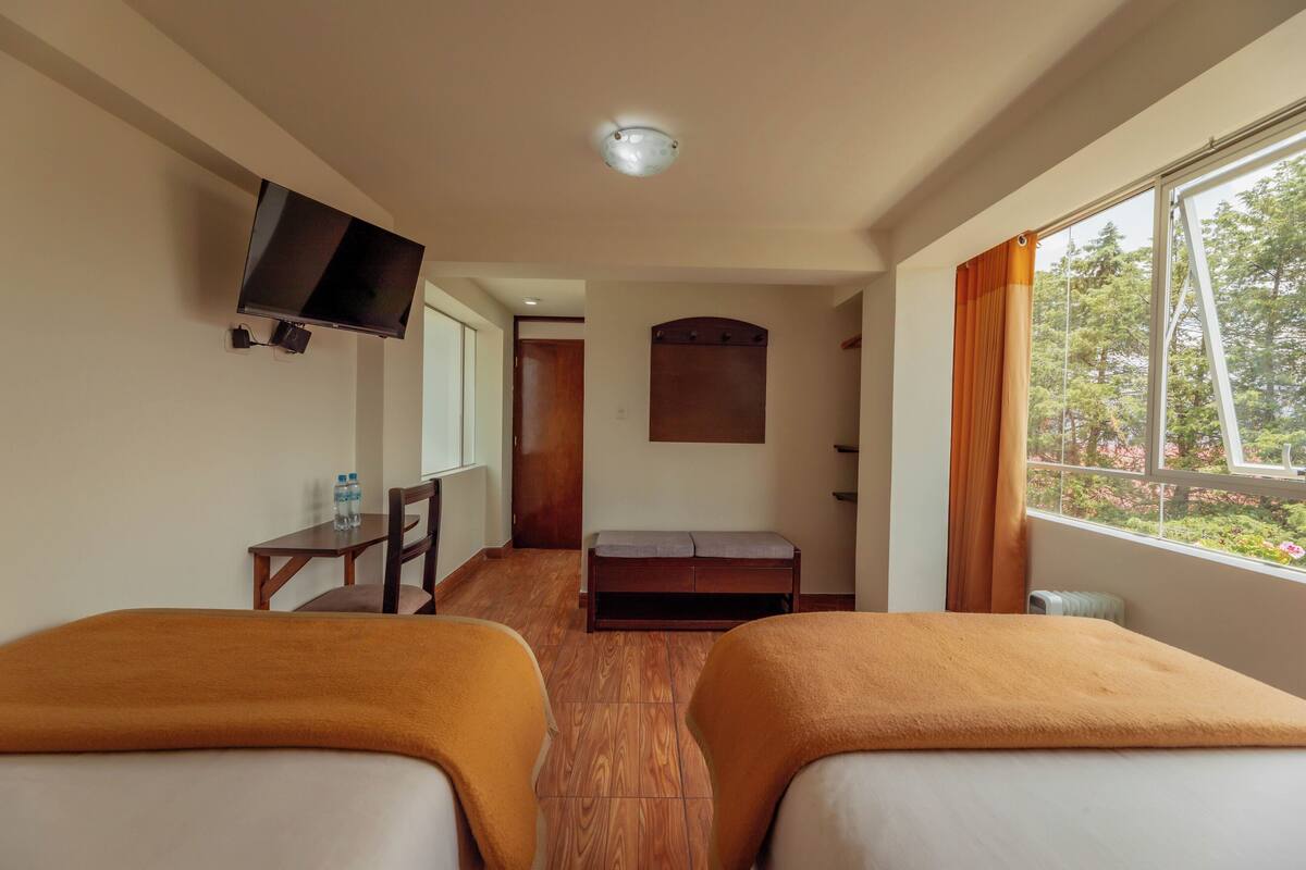 A hotel room with two beds, a TV mounted on the wall, a bench, and a window with curtains.