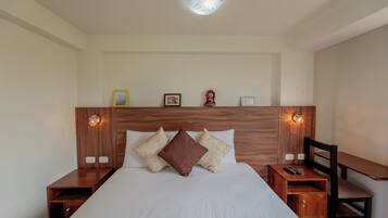 Quarto arrumado com cabeceira de madeira, cama com roupa de cama branca, duas mesas de cabeceira com luminárias e uma pequena escrivaninha com cadeira.