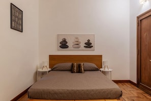 Una cama bien hecha con un cabecero de madera, dos mesitas de noche con lámparas y una obra de arte en la pared que muestra piedras apiladas.