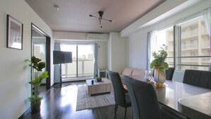 Una sala moderna con un sofá, una mesa de comedor y sillas. Hay un televisor, una planta y una ventana con vistas a edificios.