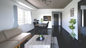 Una sala moderna con sofá, mesa de centro y comedor con sillas y mesa. Se observa una cocina al fondo, una escalera y una planta en maceta.